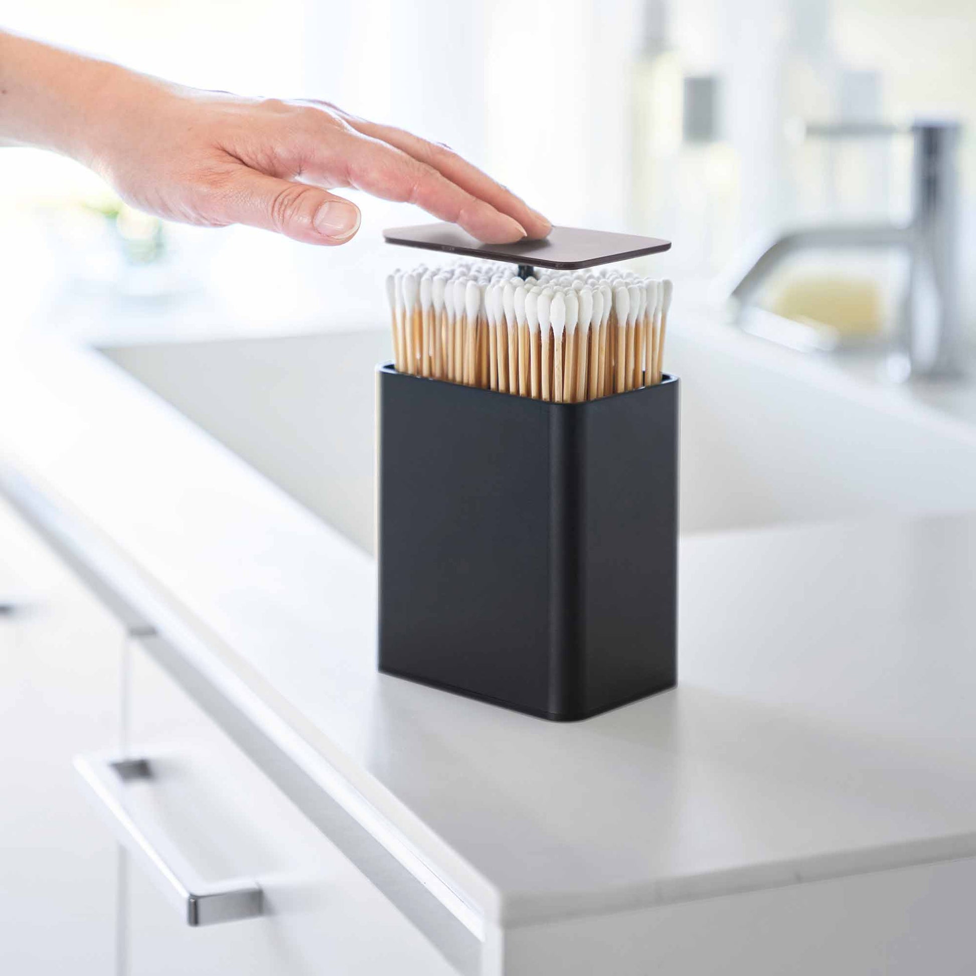 Hand pushes black cotton swab push-top case, revealing interior Qtip storage in a minimalist bathroom.