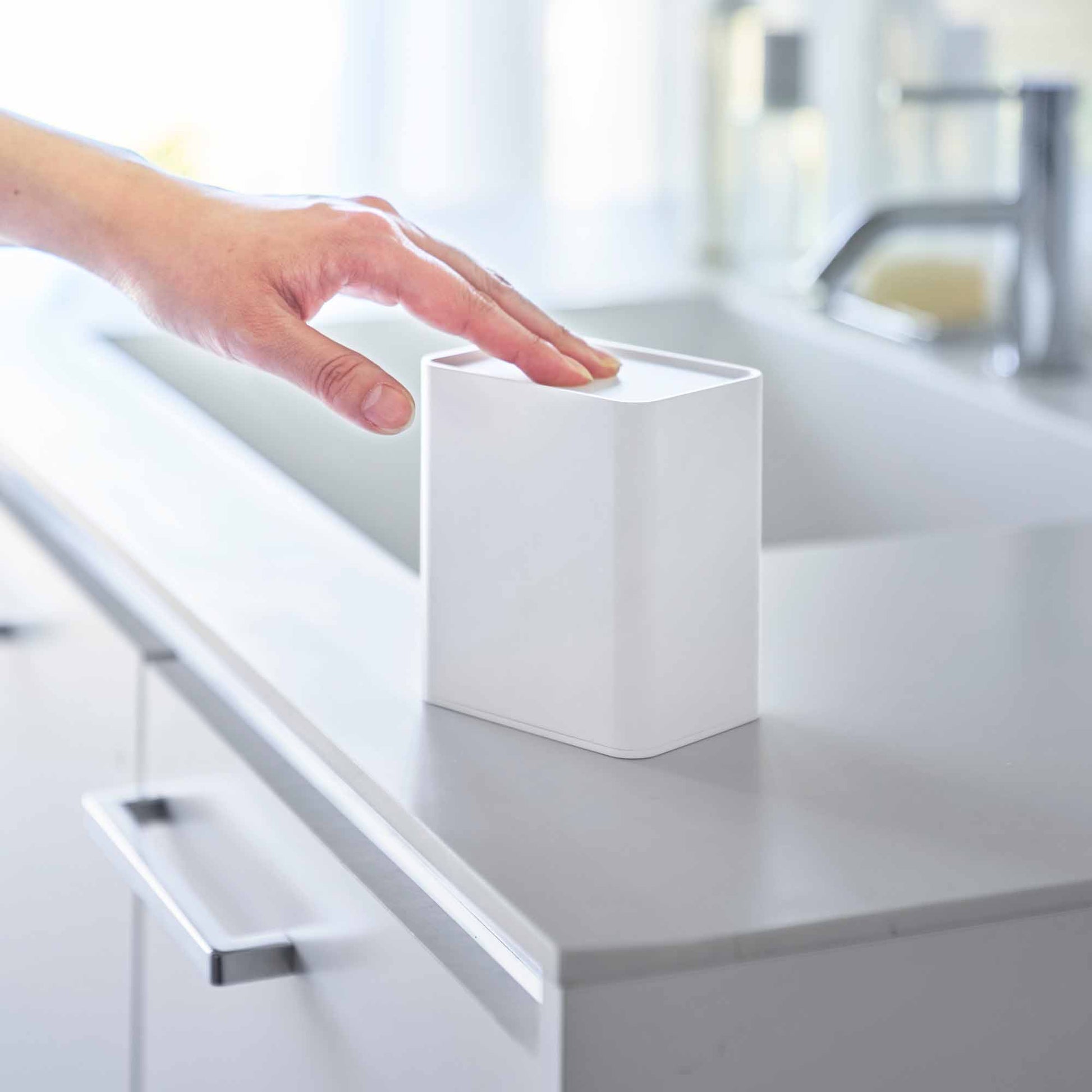 Hand pushes White cotton swab push-top case down, concealing interior Qtip storage in a minimalist bathroom.