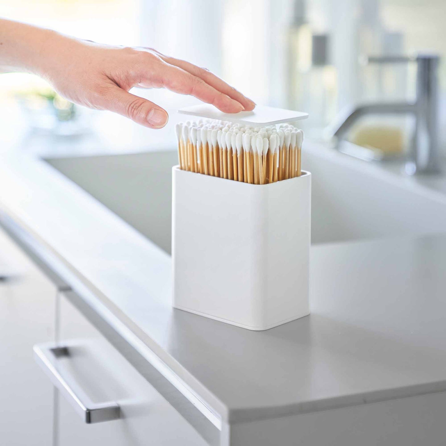 Hand pushes White cotton swab push-top case, revealing interior Qtip storage in a minimalist bathroom.
