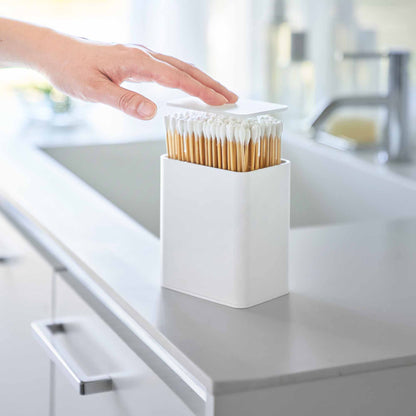 Hand pushes White cotton swab push-top case, revealing interior Qtip storage in a minimalist bathroom.