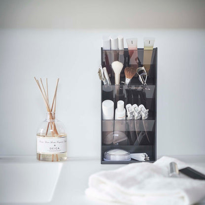 A front view of a black rectangular resin cosmetics organizer on a white bathroom counter. It has three deep adjustable black transparent trays that sit diagonally with matching dividers placed in the middle of each tray.