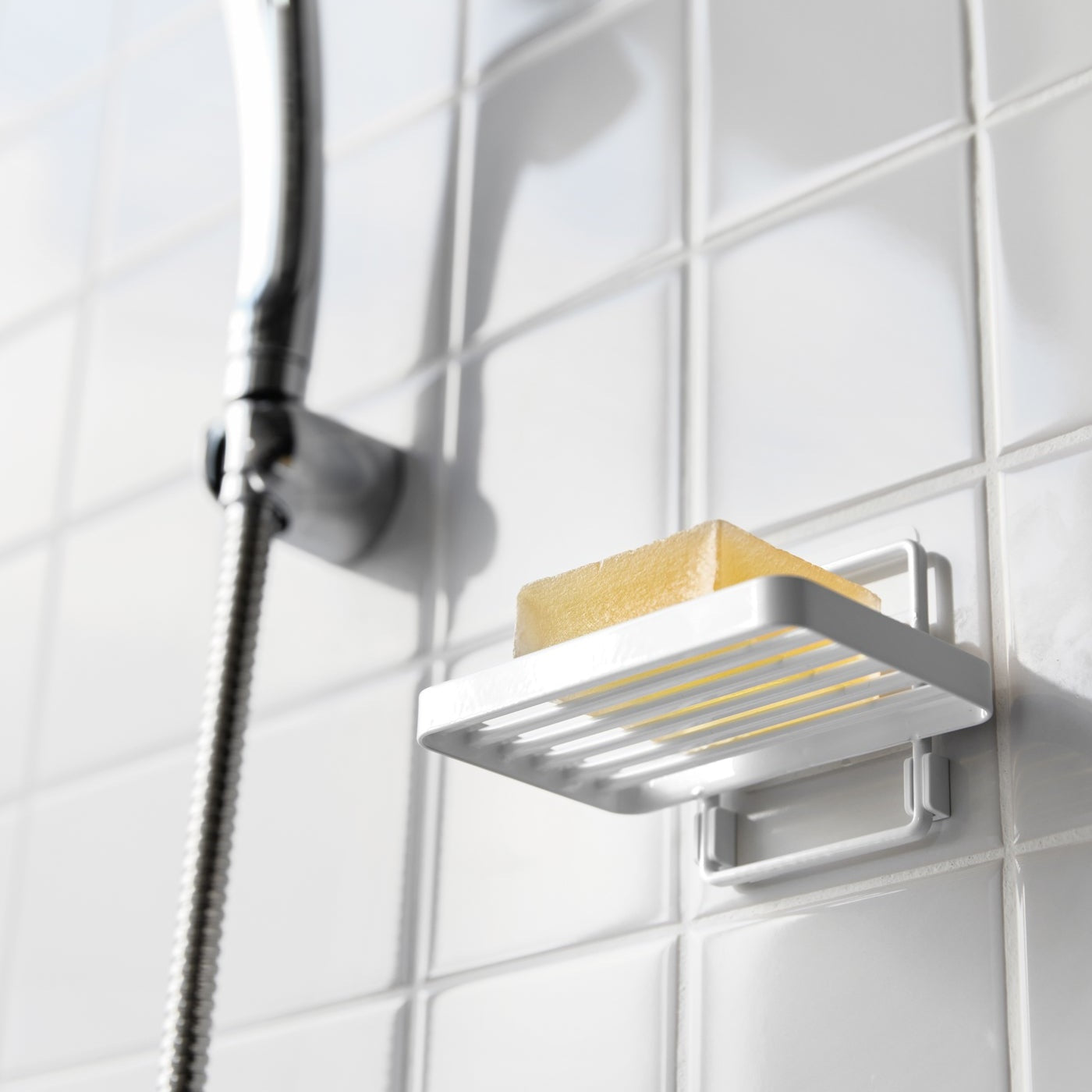 Yamazaki Home's white Traceless Adhesive Soap Tray in a shower, holding a bar of soap next to a silver shower hose on tiled wall.
