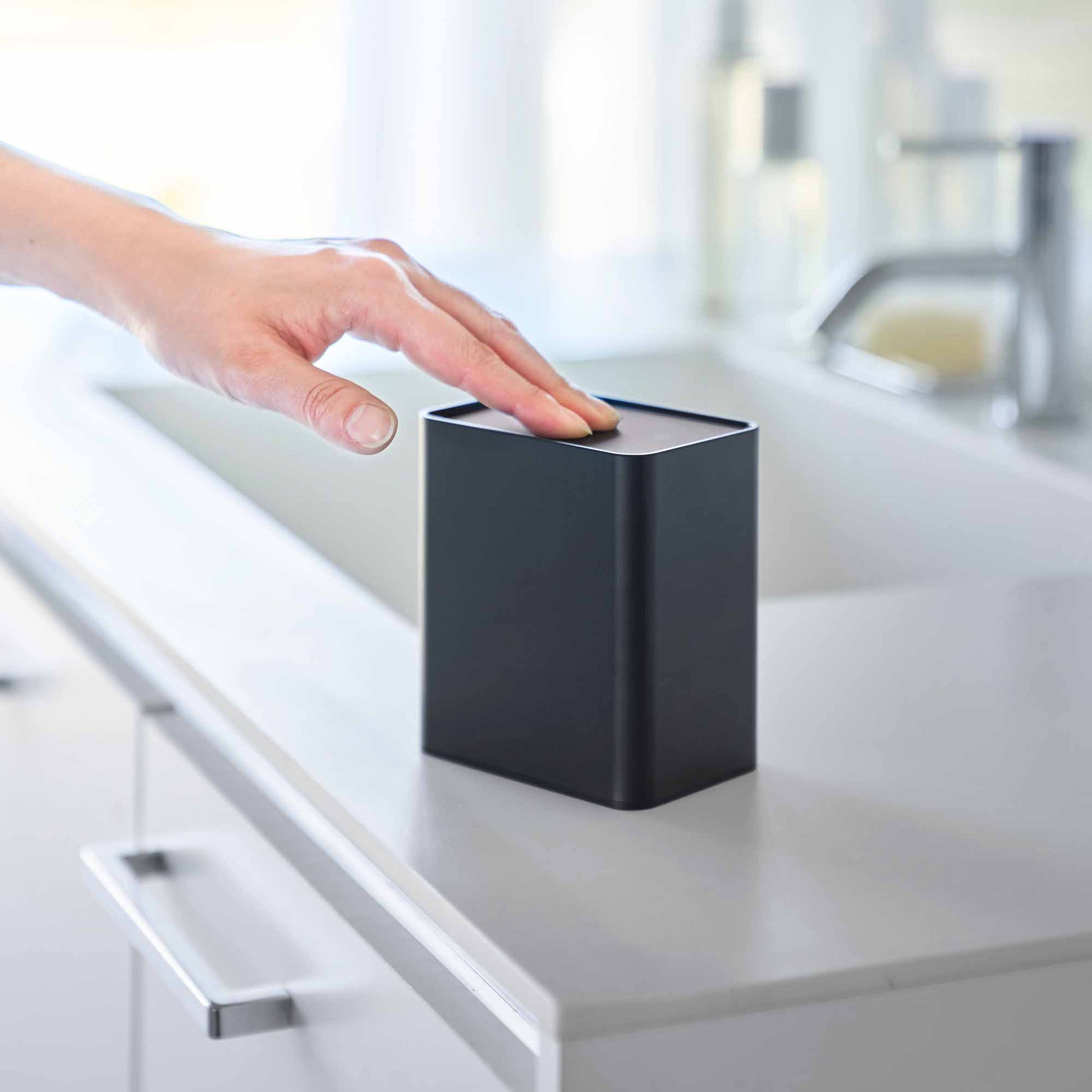 Hand pushes black cotton swab push-top case, revealing interior Qtip storage in a minimalist bathroom.