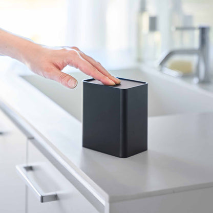 Hand pushes black cotton swab push-top case, revealing interior Qtip storage in a minimalist bathroom.