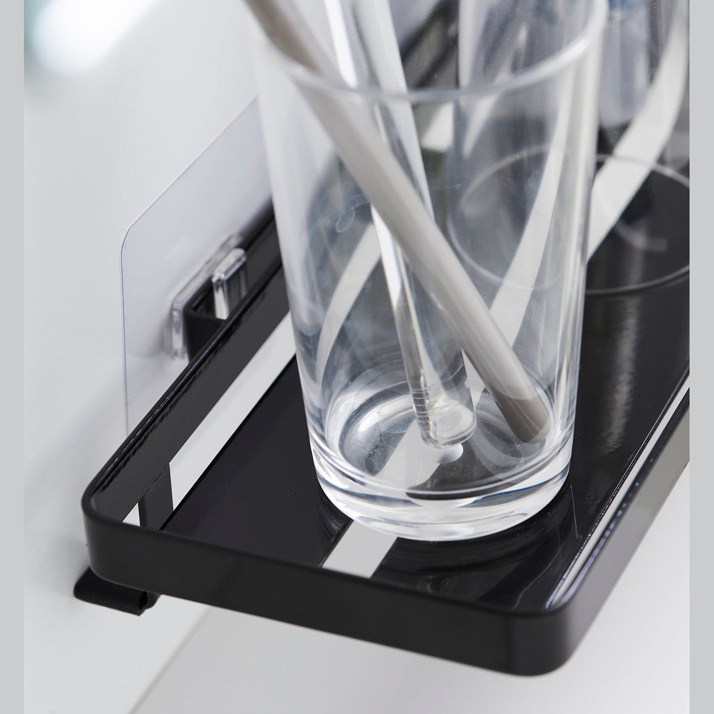 Black steel vanity tray is installed on a wall behind a sink via a translucent film hook. The tray has open slats for drainage and holds various skin care items.