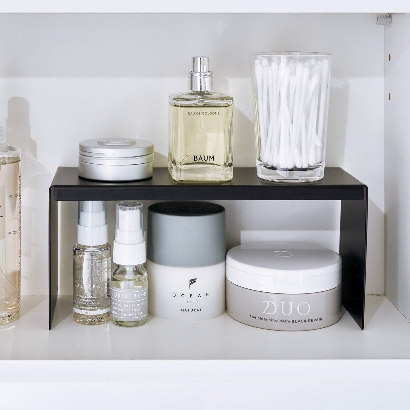 Open medicine cabinet showing black countertop riser on bottom shelf. Upper level of the riser holds perfume and hair products, while skincare and small sprays are stored on the bottom.