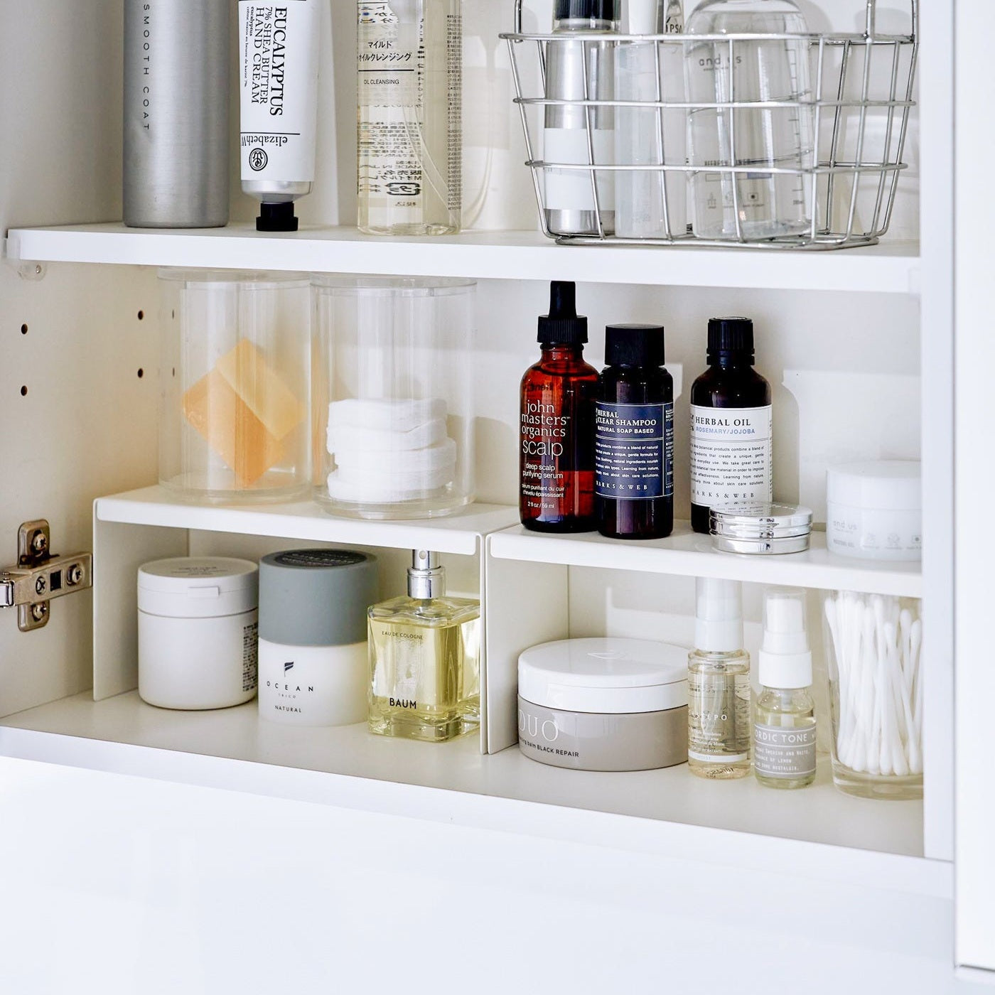 Open medicine cabinet showing two white countertop risers on bottom shelf, side by side. Upper level of the riser holds perfume and hair products, while skincare and small sprays are stored on the bottom.