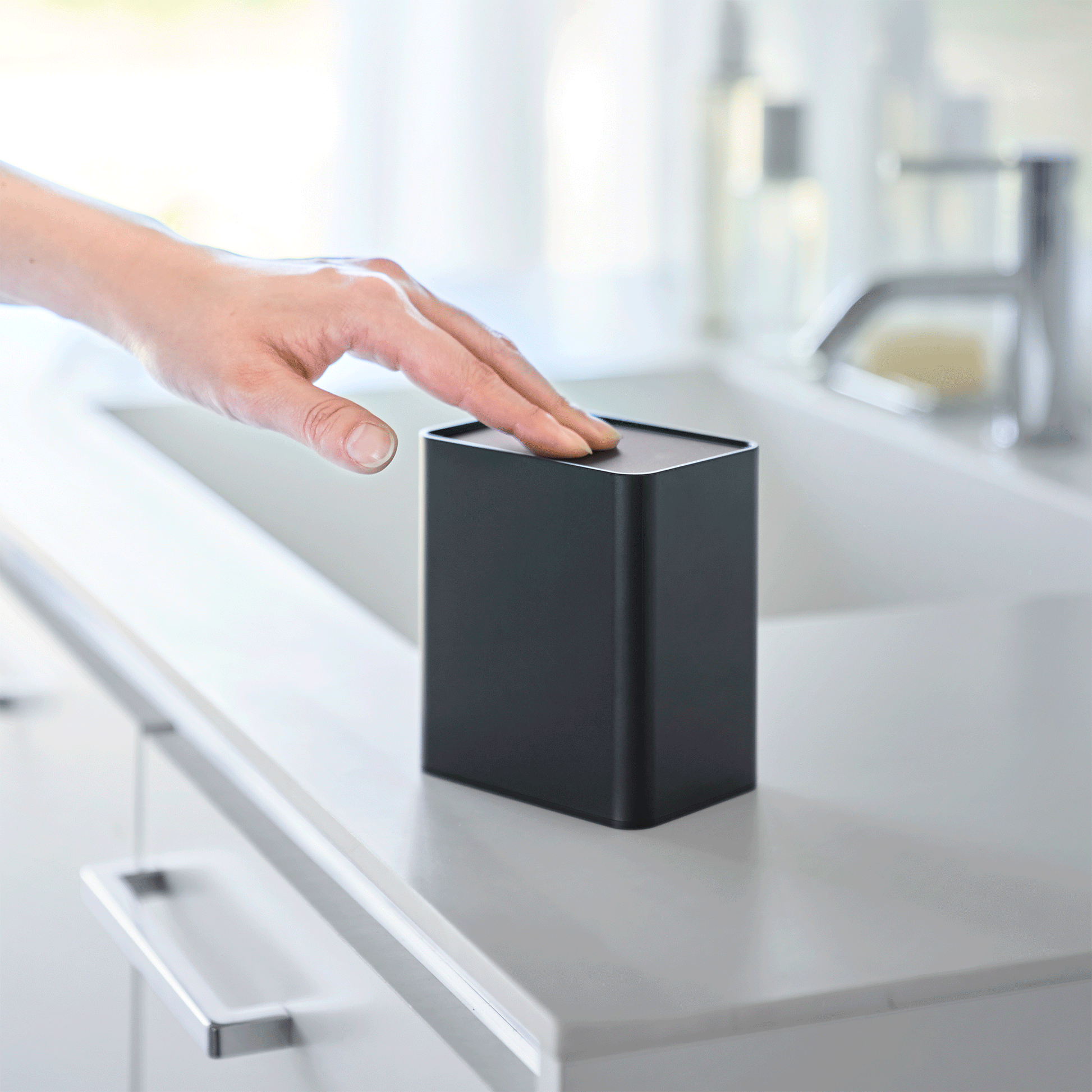 Hand pushes black cotton swab push-top case, revealing interior Qtip storage in a minimalist bathroom.