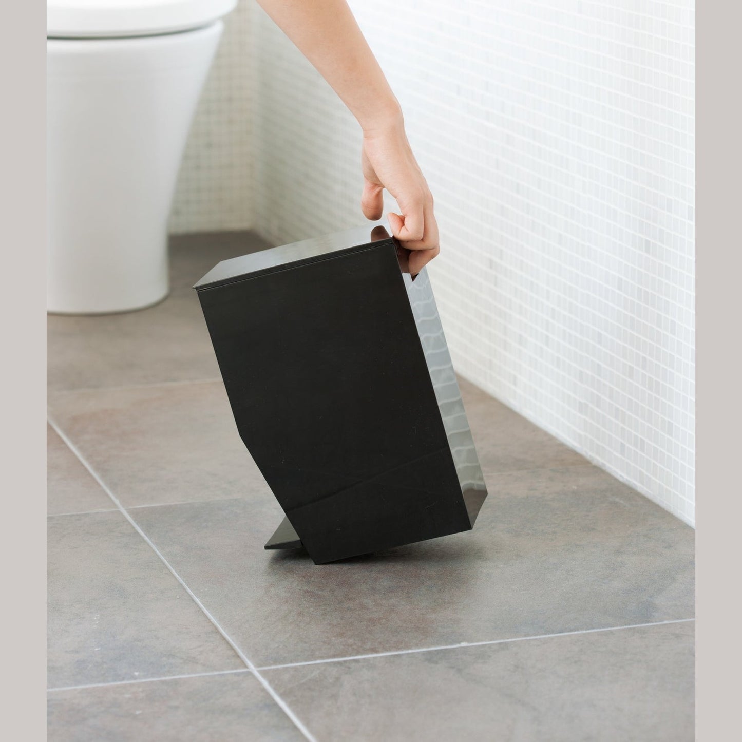Side view of black Step Trash Can held by handle in bathroom by Yamazaki Home.