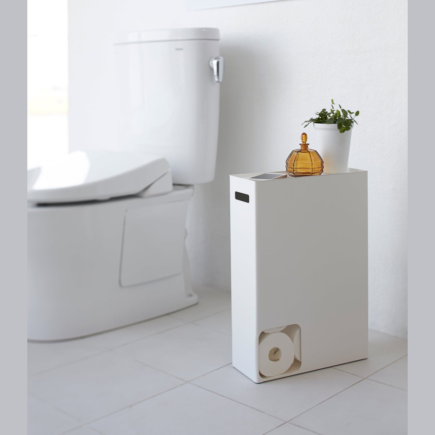 Side view of white Toilet Paper Stocker displaying phone and plant in bathroom by Yamazaki Home.