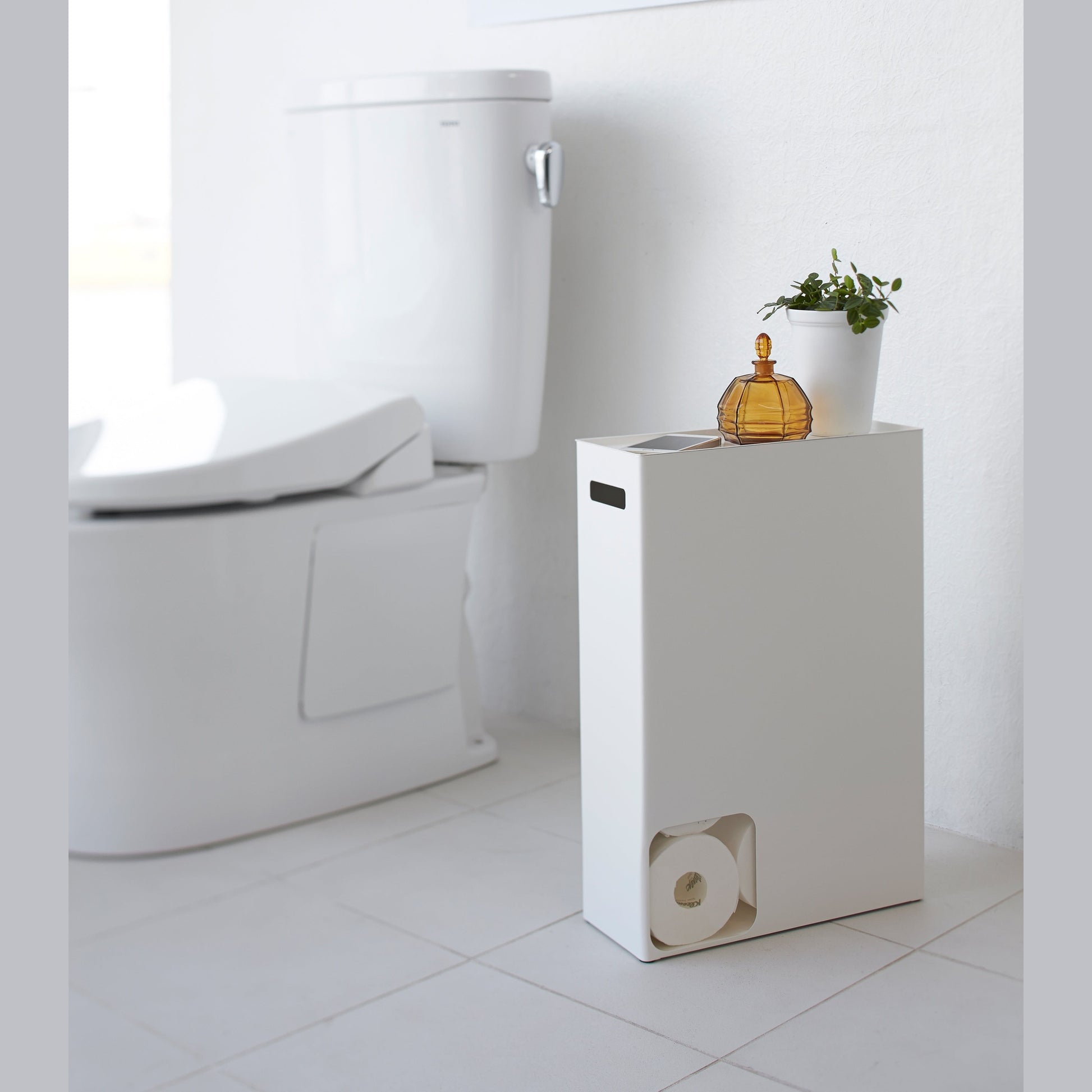 Side view of white Toilet Paper Stocker displaying phone and plant in bathroom by Yamazaki Home.