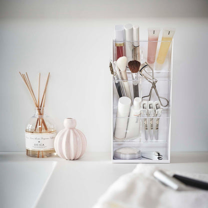 A front view of a white rectangular resin cosmetics organizer on a white bathroom counter. It has three deep transparent trays that sit diagonally with adjustable transparent dividers placed in the middle of each tray. The top tray holds lipsticks and glosses