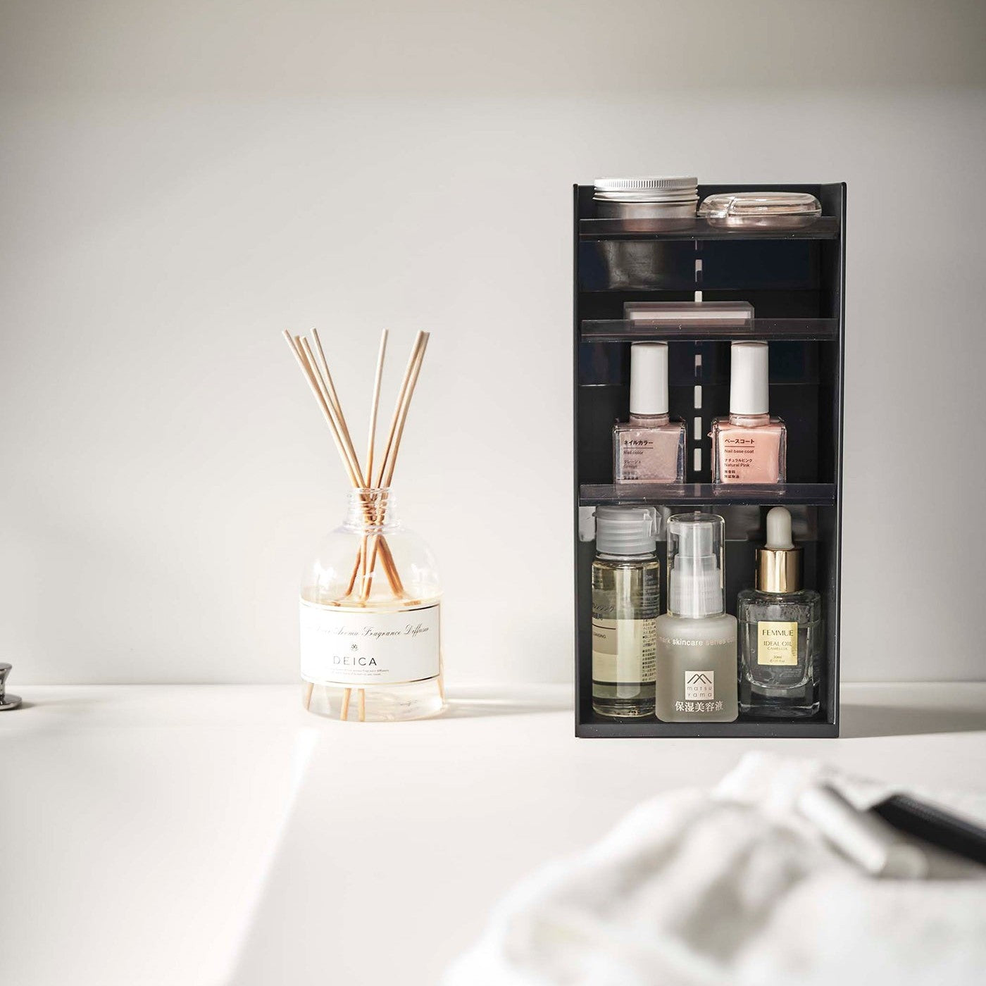 A black resin rectangular cosmetics organizer sits on a white bathroom counter. The organizer has three transparent shelves with upward facing lips to prevent products from falling-out.
