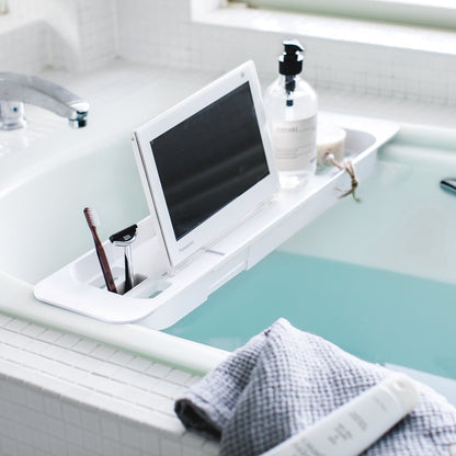 White Expandable Bathtub Caddy holding tablet and cleaning products in bathroom by Yamazaki Home.