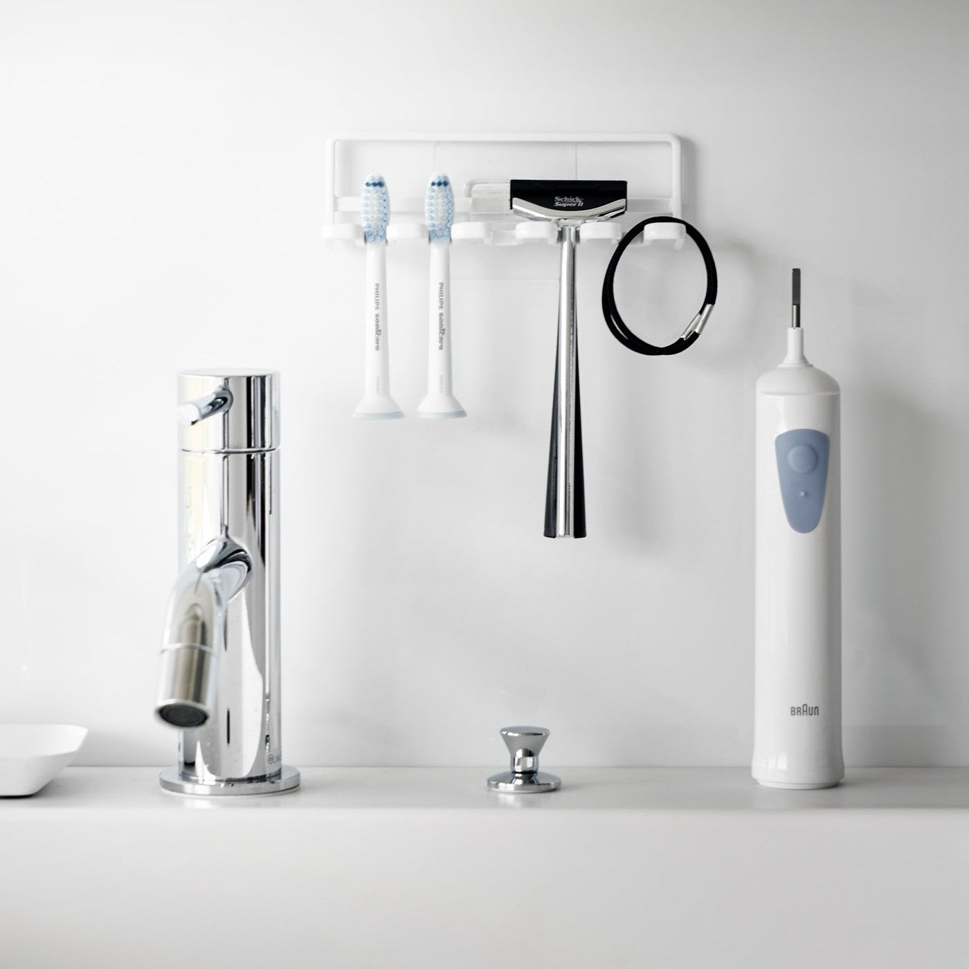 Yamazaki Home's white Traceless Adhesive Toothbrush Holder on a bathroom wall, holding two toothbrushes and a razor beside a faucet.