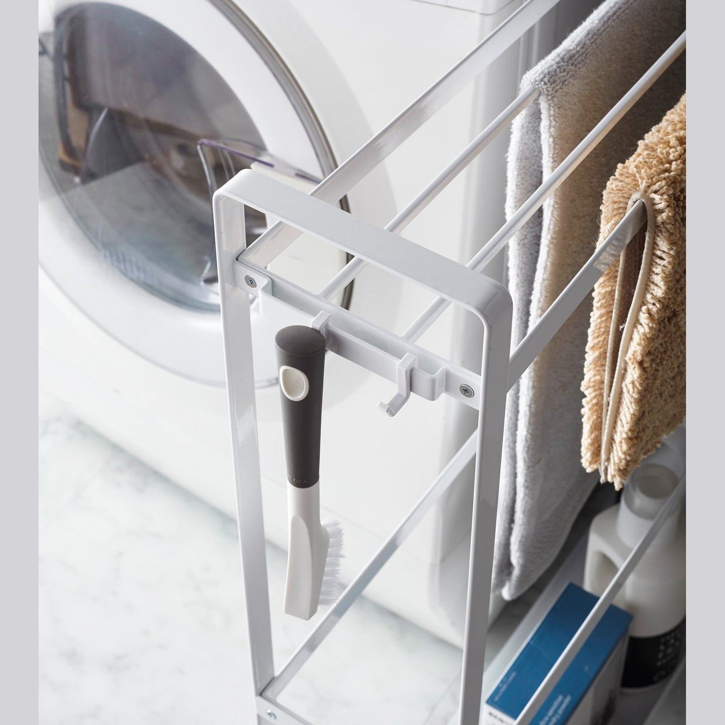 Close up corner view of white Rolling Towel Rack holding cleaning items and mats in laundry room by Yamazaki Home.