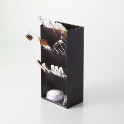 A black resin rectangular cosmetics organizer with an open face and top sits on a white surface. A light is shining at the product from the upper right. It has three deep black transparent trays that sit diagonally with matching dividers placed in the middle of each tray. The bottom of the organizer has a slightly raised lip and acts as additional storage. Various cosmetics are stored in each tray and the bottom of the organizer holds nail clippers and a circular cosmetic product.