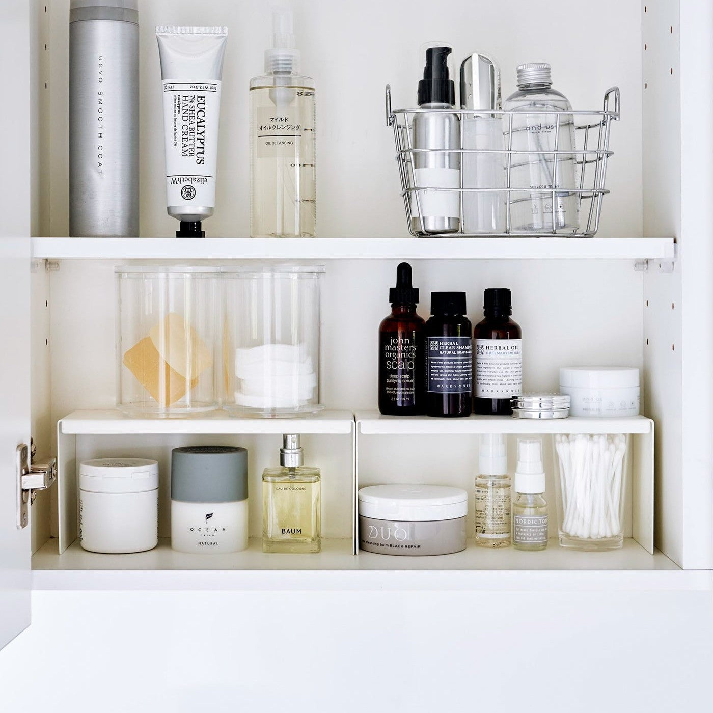 Open medicine cabinet showing two white countertop risers on bottom shelf, side by side. Upper level of the riser holds perfume and hair products, while skincare and small sprays are stored on the bottom.