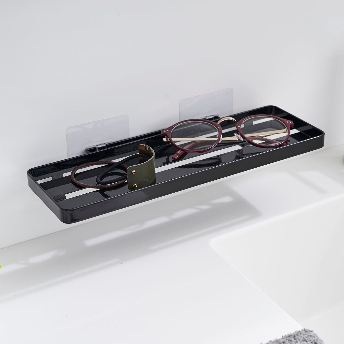 Black steel vanity tray is installed on a wall behind a sink via a translucent film hook. The tray has open slats for drainage and holds hair ties and reading glasses.