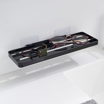 Black steel vanity tray is installed on a wall behind a sink via a translucent film hook. The tray has open slats for drainage and holds hair ties and reading glasses.