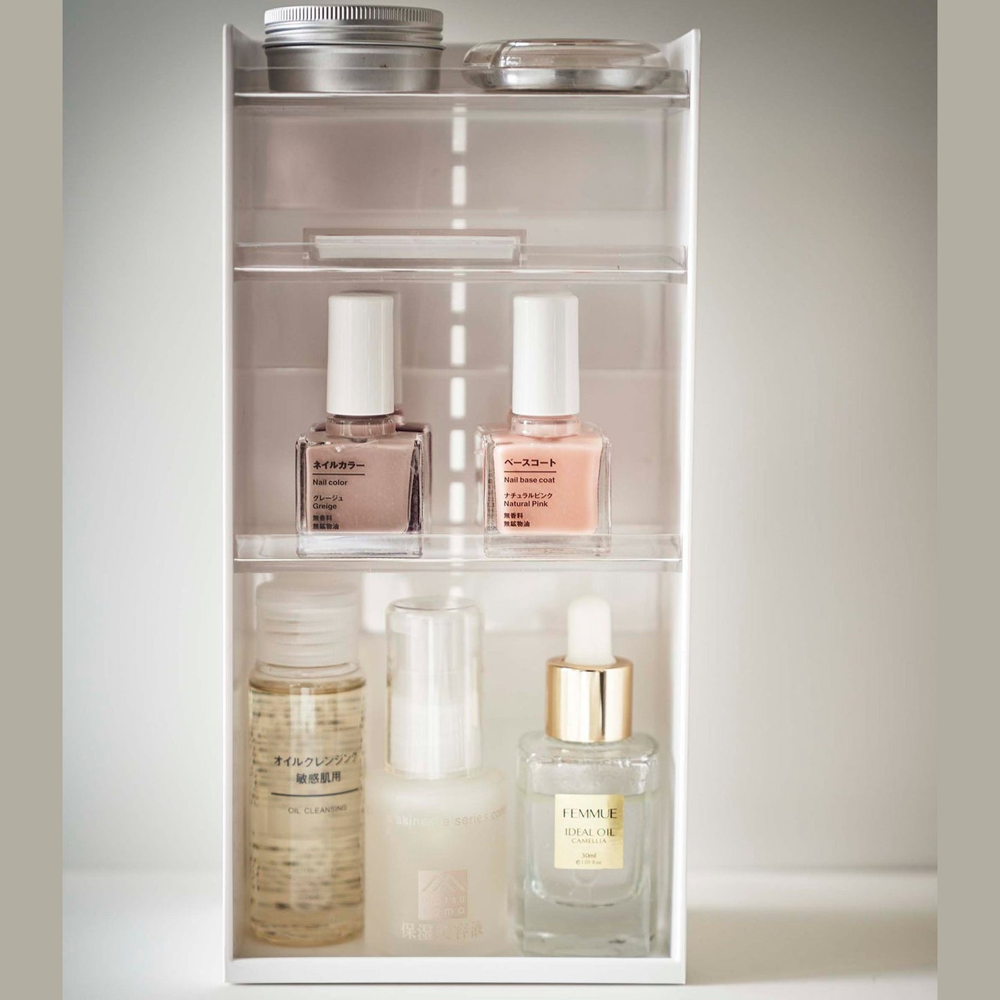On top of a white surface is a white resin rectangular cosmetics organizer with an open face and top. It has three transparent shelves with upward facing lips to prevent products from falling-out.