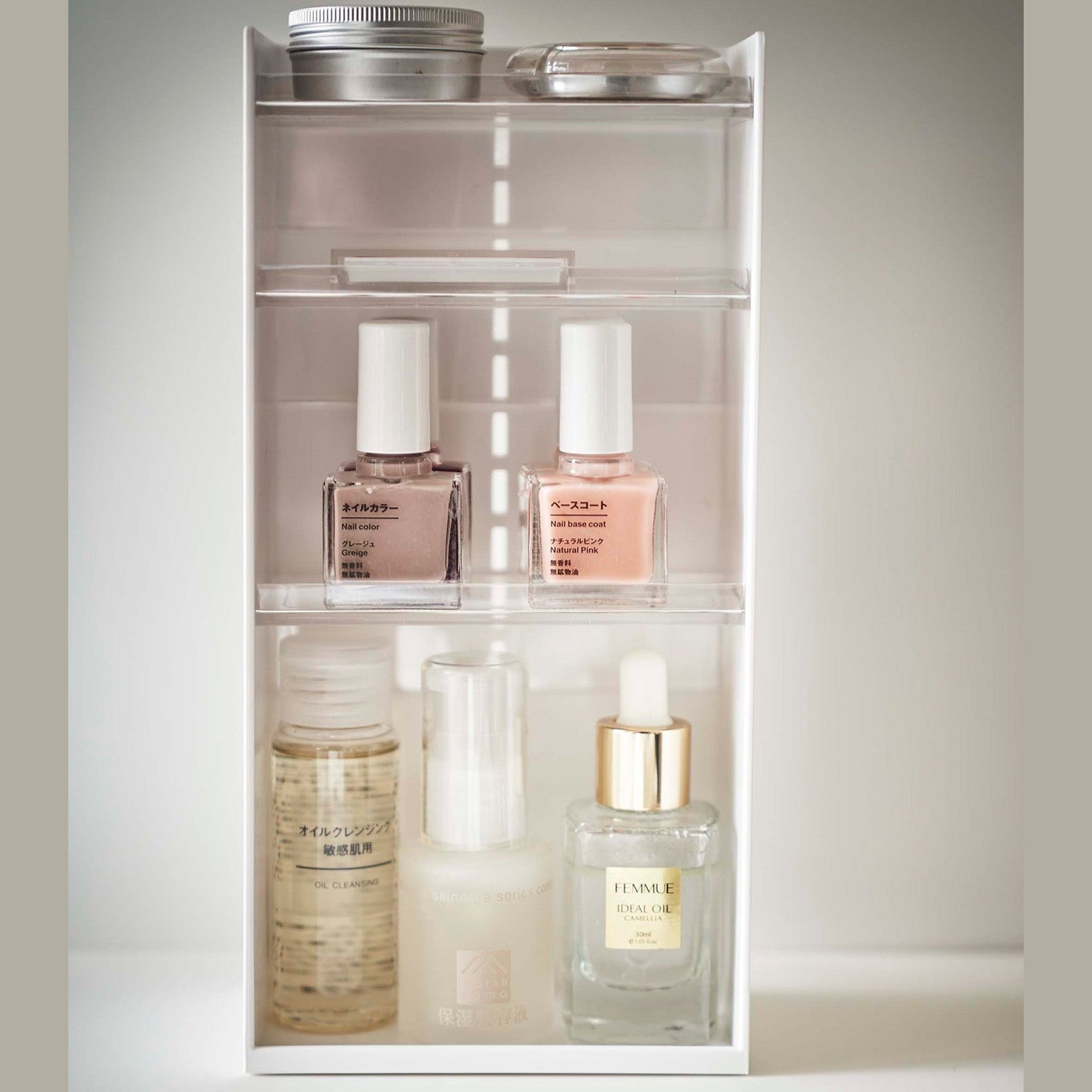 On top of a white surface is a white resin rectangular cosmetics organizer with an open face and top. It has three transparent shelves with upward facing lips to prevent products from falling-out.