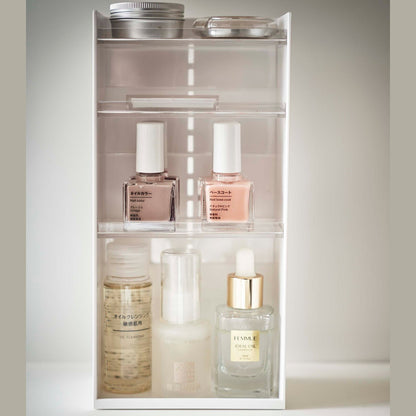 On top of a white surface is a white resin rectangular cosmetics organizer with an open face and top. It has three transparent shelves with upward facing lips to prevent products from falling-out.