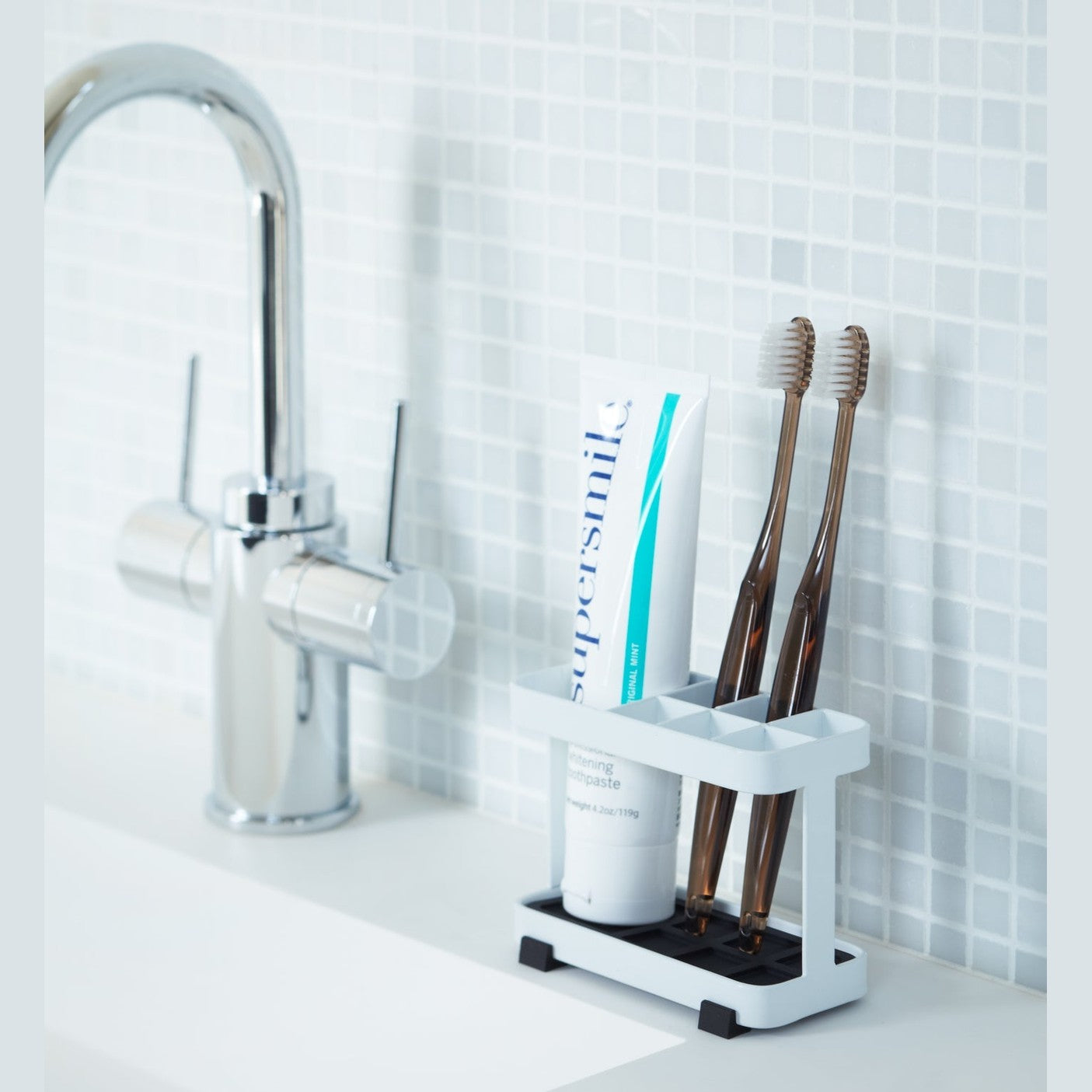 White Toothbrush Stand displaying toothpaste and toothbrushes on bathroom sink counter by Yamazaki Home.