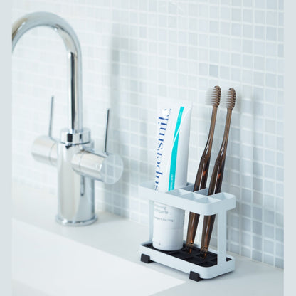 White Toothbrush Stand displaying toothpaste and toothbrushes on bathroom sink counter by Yamazaki Home.