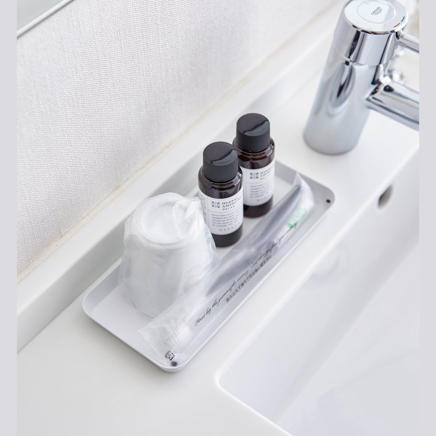 Aerial view of flat white Accessory Tray holding soap, cup, and toothbrush on bathroom sink counter by Yamazaki Home.