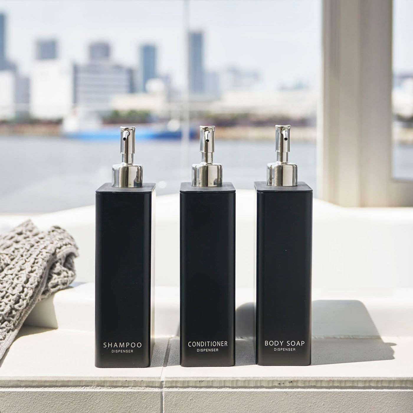 Front view of Yamazaki Home black Shampoo, Conditioner, and Body Soap dispensers on the ledge of a modern bathtub.