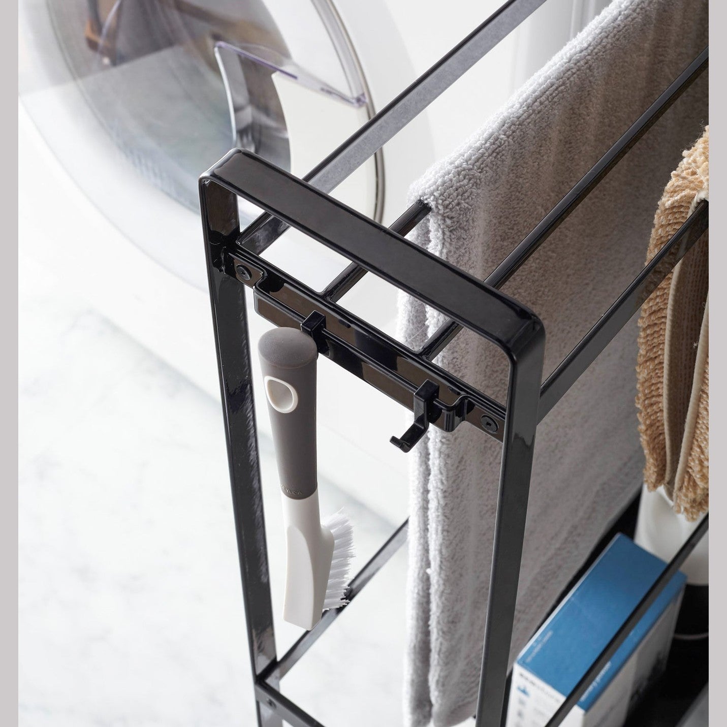 Close up corner view of black Rolling Towel Rack holding towel, mat, and cleaning items in laundry room by Yamazaki Home.