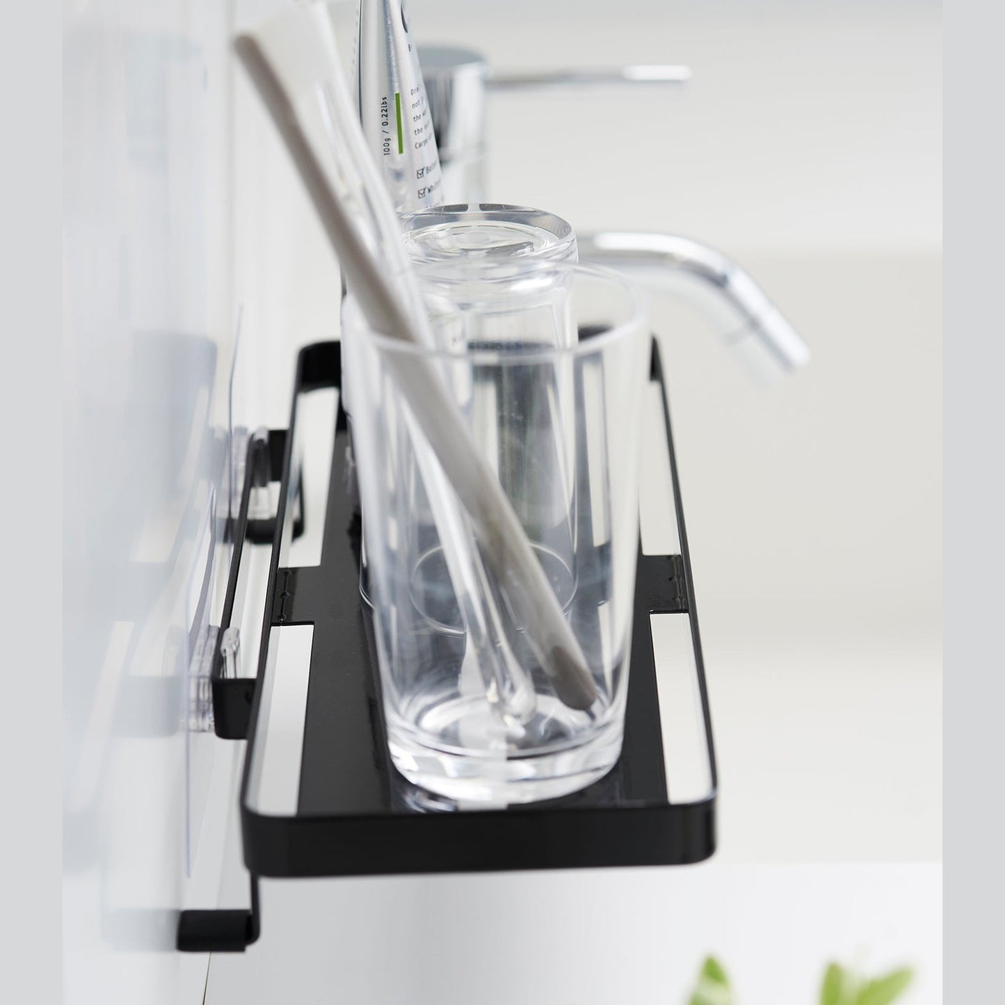 Black steel vanity tray is installed on a wall behind a sink via a translucent film hook. The tray has open slats for drainage and holds a toothbrush.