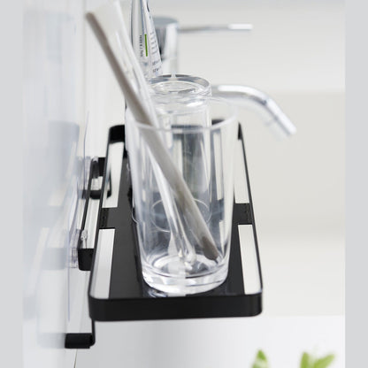 Black steel vanity tray is installed on a wall behind a sink via a translucent film hook. The tray has open slats for drainage and holds a toothbrush.