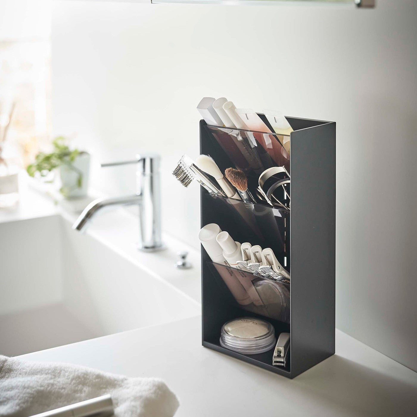 An angled view of a black rectangular resin cosmetics organizer on a white bathroom counter. It has an open face and top and three deep black adjustable transparent trays that sit diagonally with matching dividers placed in the middle of each tray. The bottom of the organizer has a raised lip and acts as additional storage.