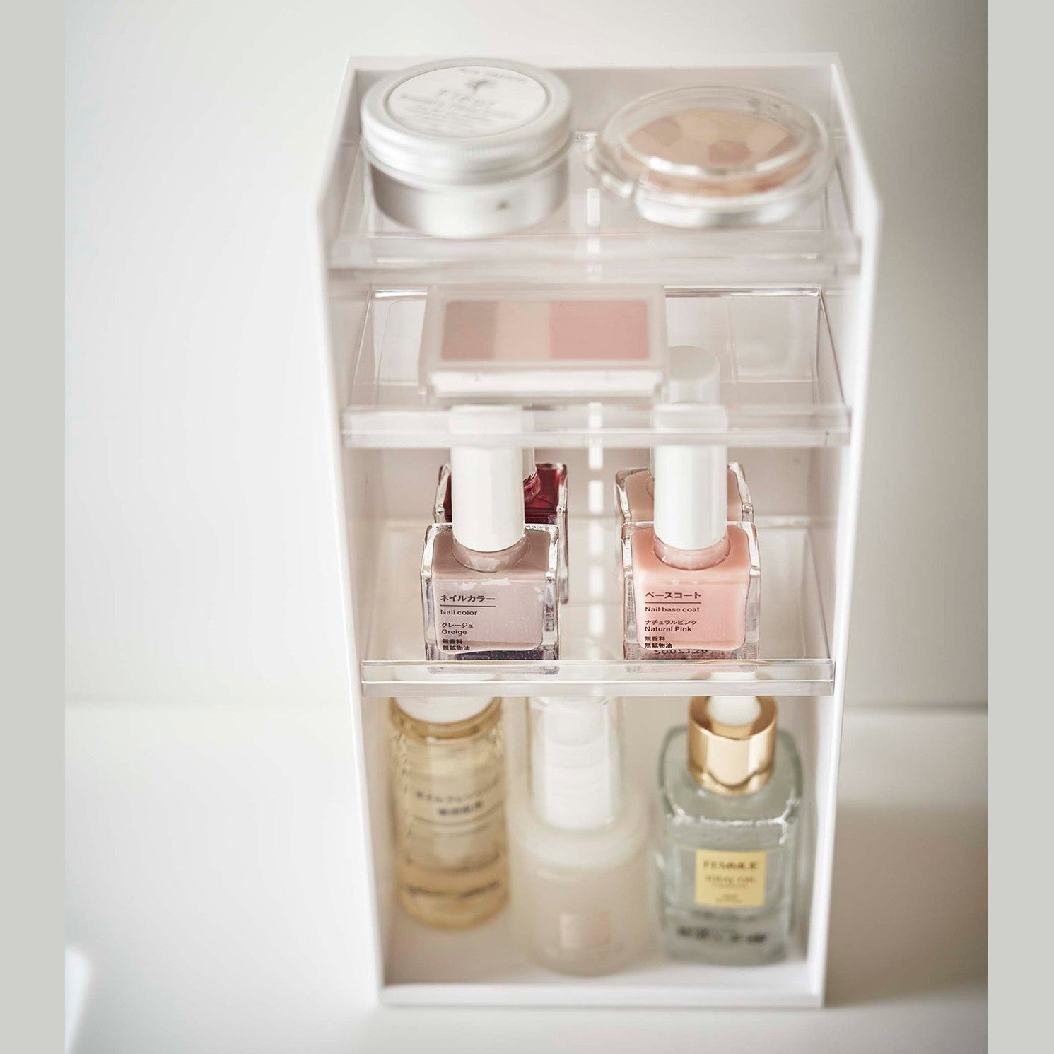A detailed upper view of a white rectangular cosmetics organizer sitting. The organizer has three transparent shelves with upward facing lips to prevent the products from falling-out.