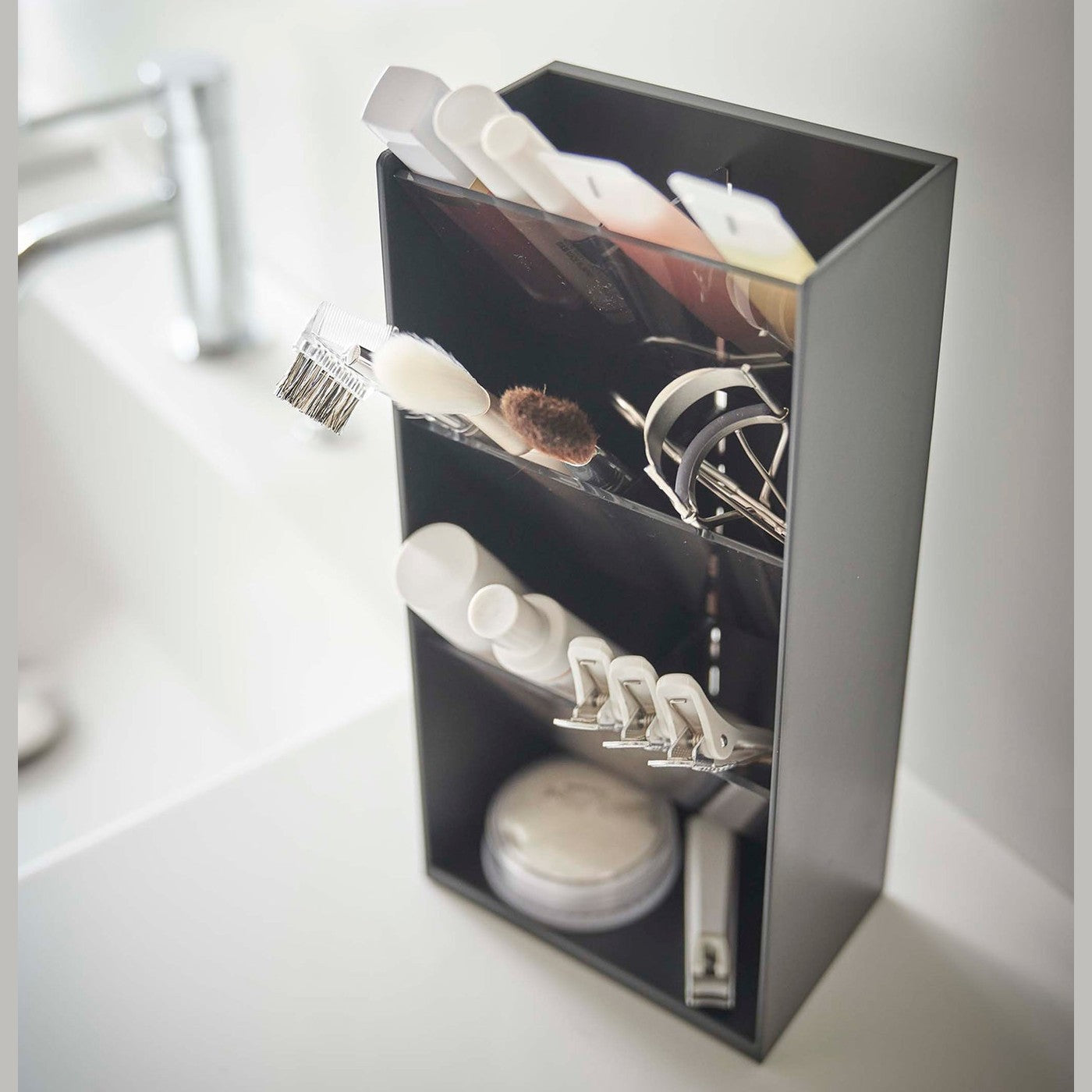 An upper view of a black rectangular resin cosmetics organizer on a white bathroom counter. It has three deep black transparent trays that lean forward diagonally with matching dividers placed in the middle of each tray. The middle tray is in focus and holds a set of cosmetic brushes and an eyelash curler. In the background is an out-of-focus sink faucet and basin.
