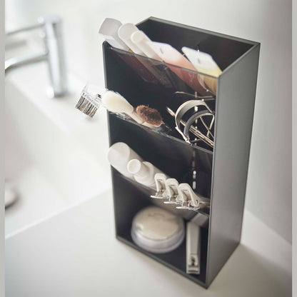 An upper view of a black rectangular resin cosmetics organizer on a white bathroom counter. It has three deep black transparent trays that lean forward diagonally with matching dividers placed in the middle of each tray. The middle tray is in focus and holds a set of cosmetic brushes and an eyelash curler. In the background is an out-of-focus sink faucet and basin.