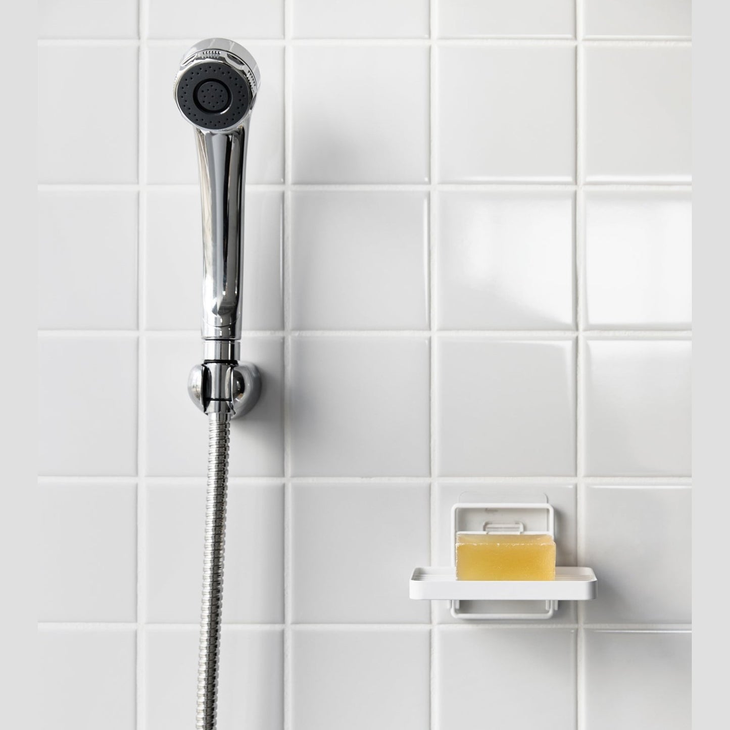 Yamazaki Home's white Traceless Adhesive Soap Tray in a shower, holding a bar of soap next to a silver shower hose on tiled wall.