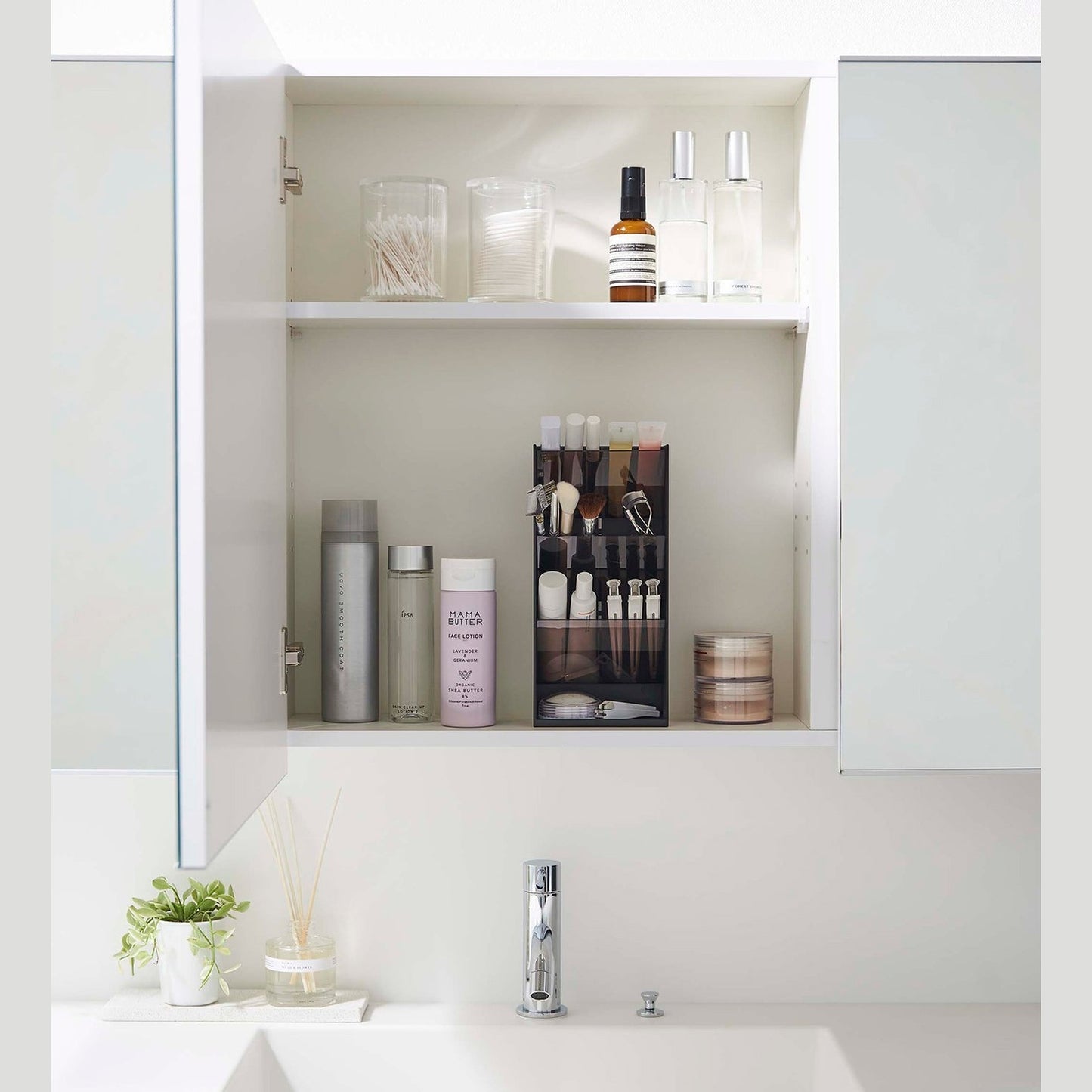 A front-facing view of an opened white medicine cabinet. Sunlight is focused on the right upper corner. The top half of the cabinet holds face serums and two clear containers with cotton swabs and rounds.