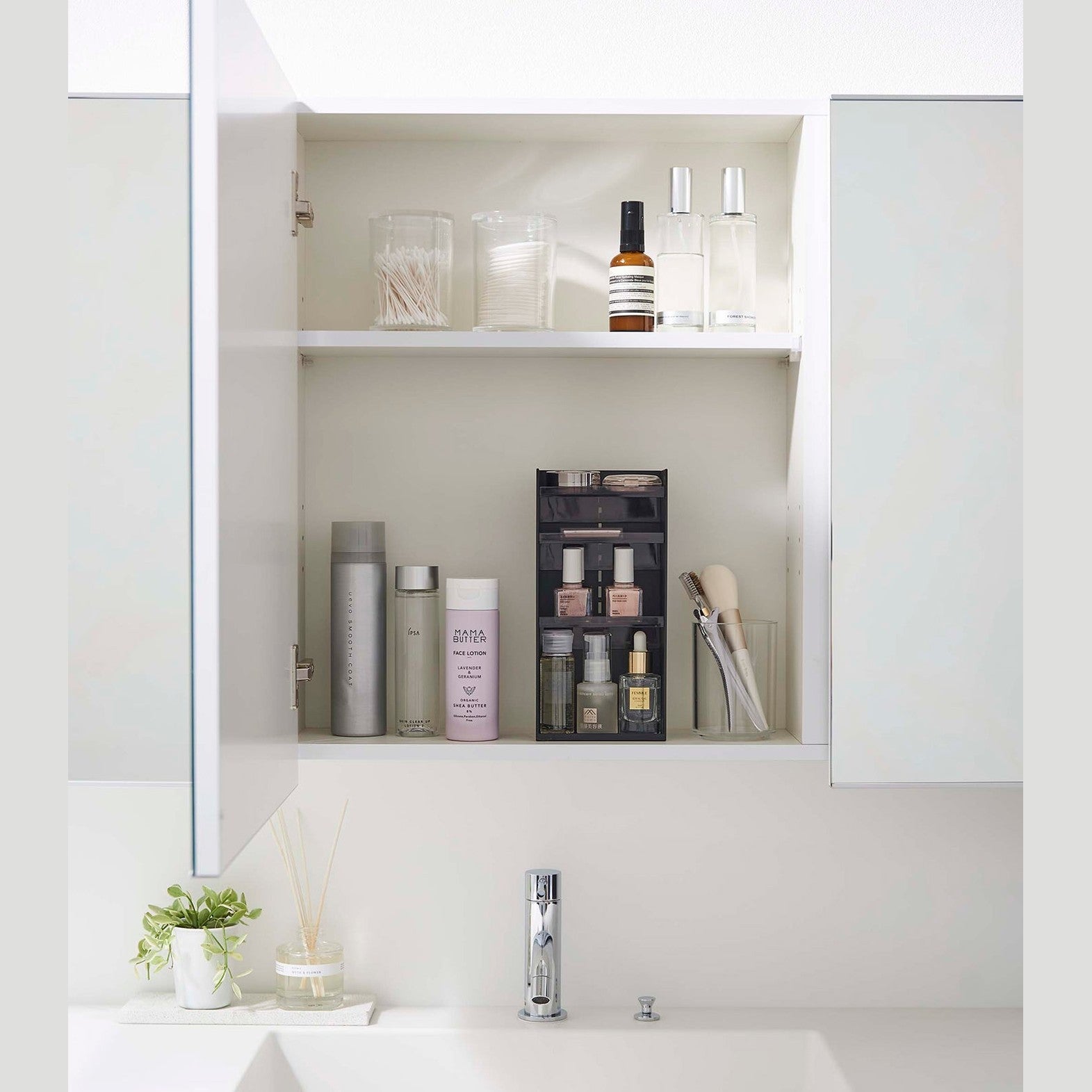 A white medicine cabinet is open to display the inside contents. Sunlight is focused on the right upper corner. Below is a bathroom sink. On the sinks ledge is a plant and oil diffuser.