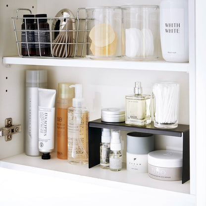 Open medicine cabinet showing black countertop riser on bottom shelf. Upper level of the riser holds perfume and hair products, while skincare and small sprays are stored on the bottom.