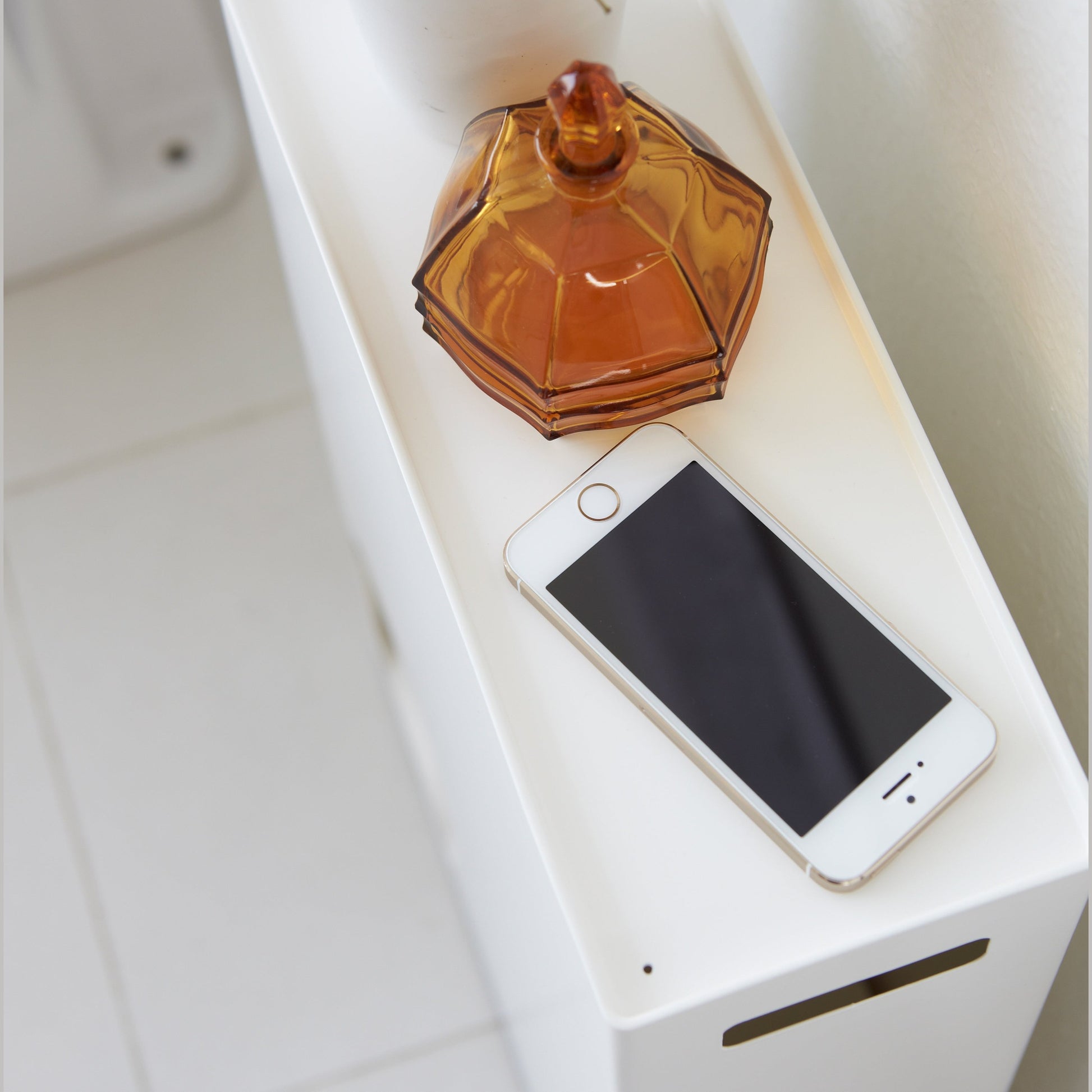 Close up aerial view of white Toilet Paper Stocker top holding plant, jar, and phone in bathroom by Yamazaki Home.