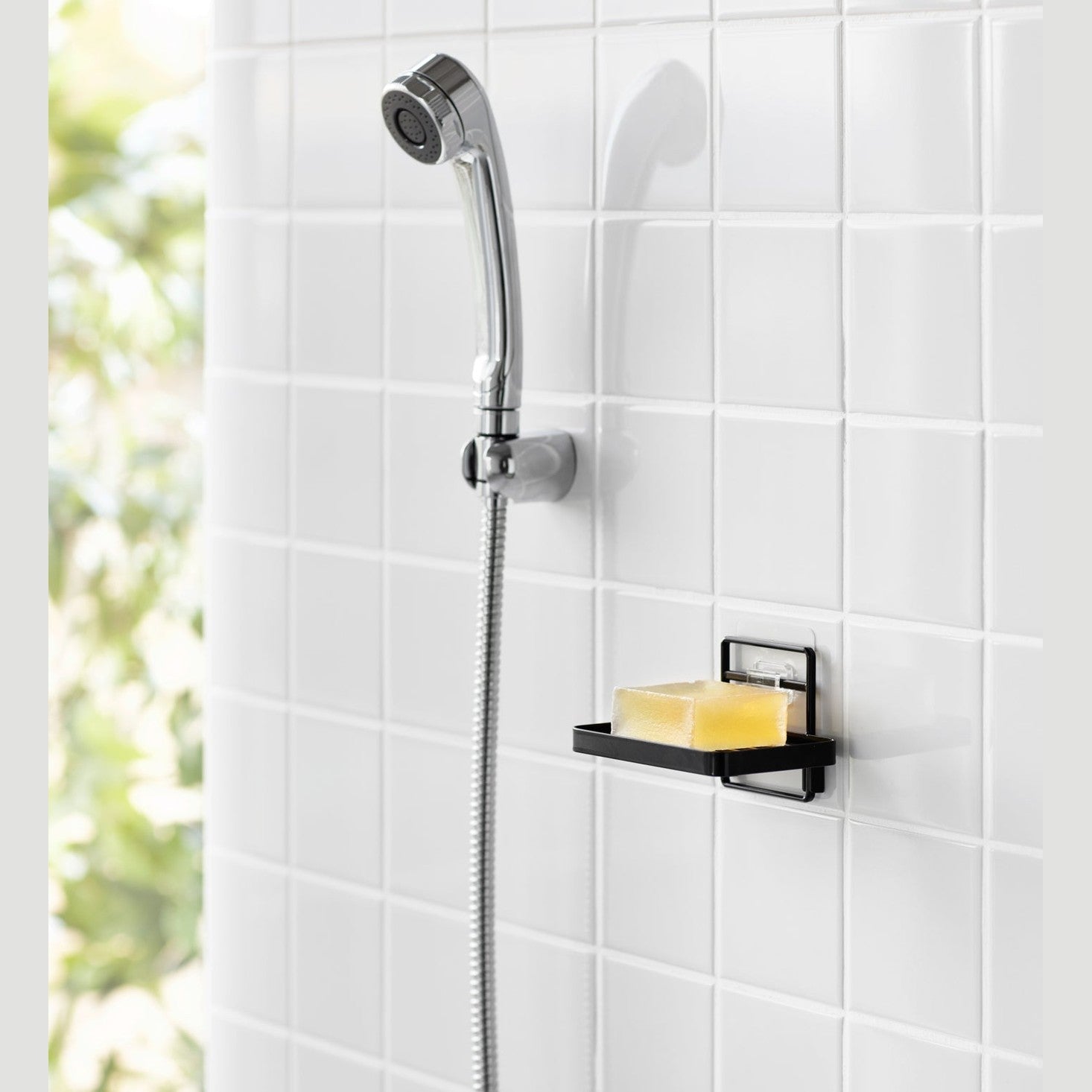 Yamazaki Home's black Traceless Adhesive Soap Tray in a shower, holding a bar of soap next to a silver shower hose on tiled wall.