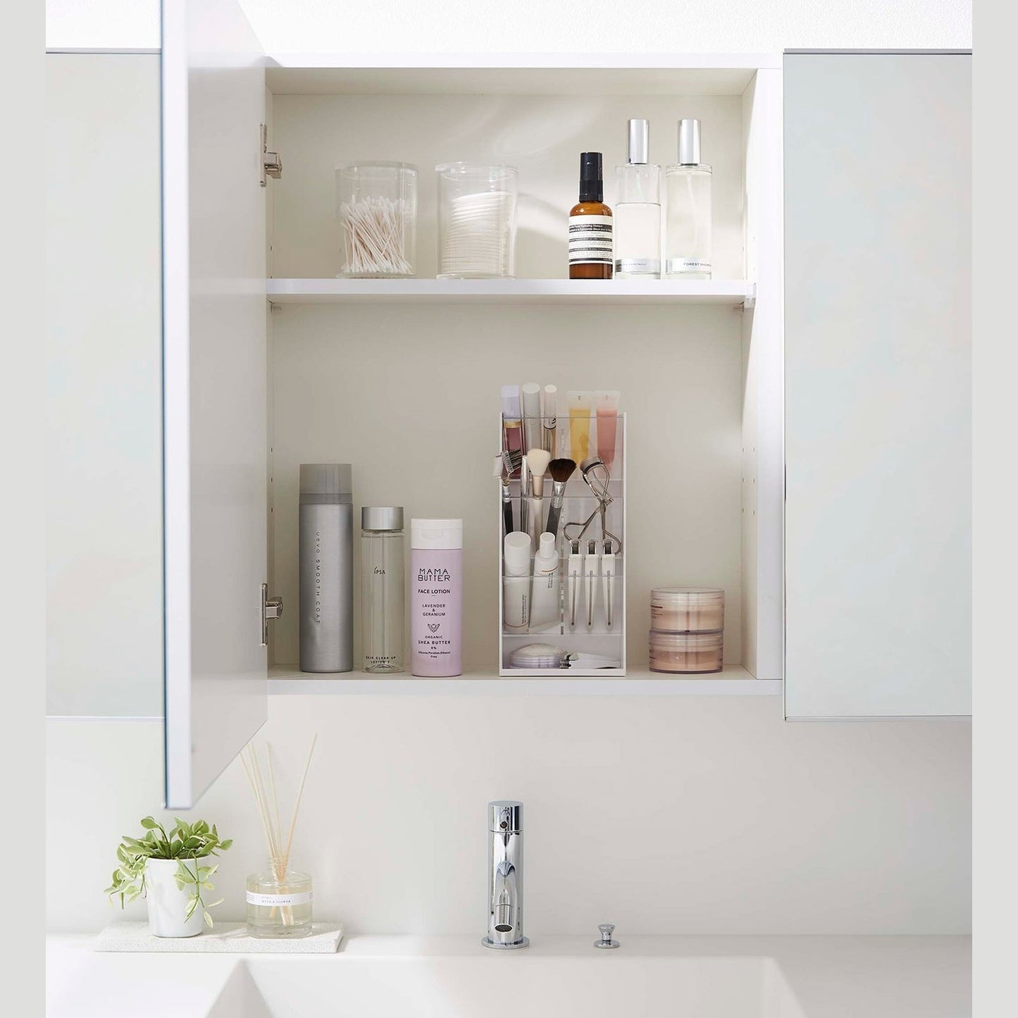 A direct view of an opened white medicine cabinet. Sunlight is focused on the right upper corner. The top half of the cabinet holds face serums and two clear containers with cotton swabs and rounds.