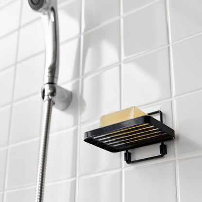 Yamazaki Home's black Traceless Adhesive Soap Tray in a shower, holding a bar of soap next to a silver shower hose on tiled wall.