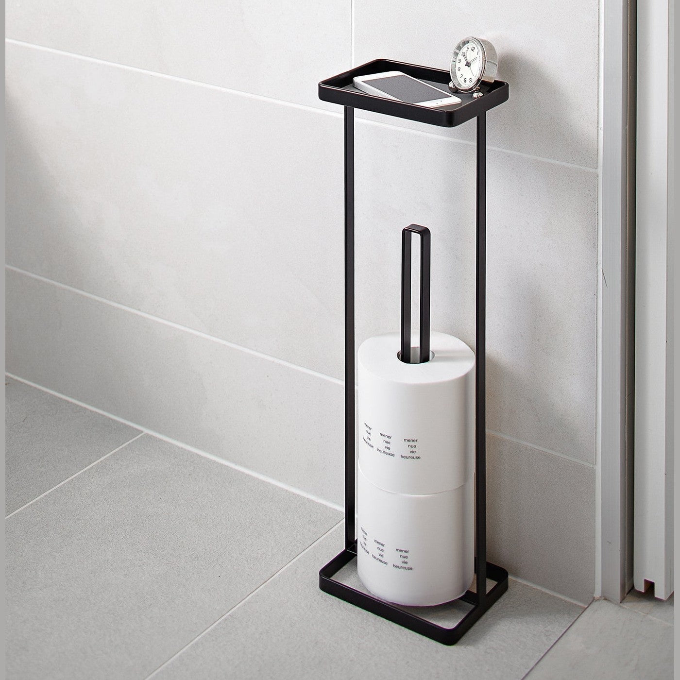Black Toilet Paper Stocker holding phone, clock and toilet paper in bathroom by Yamazaki home.