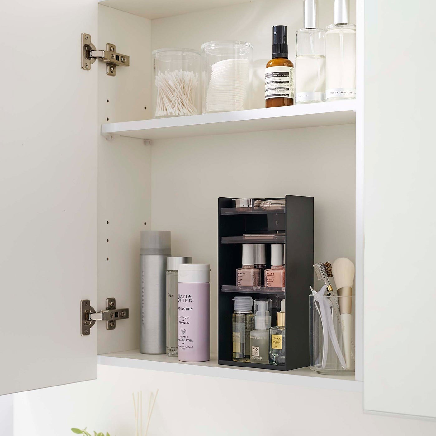A white medicine cabinet is open to display the inside contents. Sunlight is focused on the right upper corner. Below is a bathroom sink. On the sinks ledge is a plant and oil diffuser. A white shelf divides the cabinet. The bottom section features a cosmetic organizer.