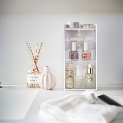 A white rectangular cosmetics organizer sits on a white bathroom counter. The organizer has three transparent shelves with upward facing lips to prevent products from falling-out.