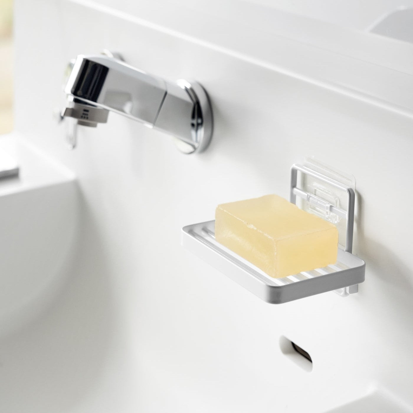 Yamazaki Home's white Traceless Adhesive Soap Tray mounted on a bathroom wall, holding a bar of yellow soap next to a chrome faucet.